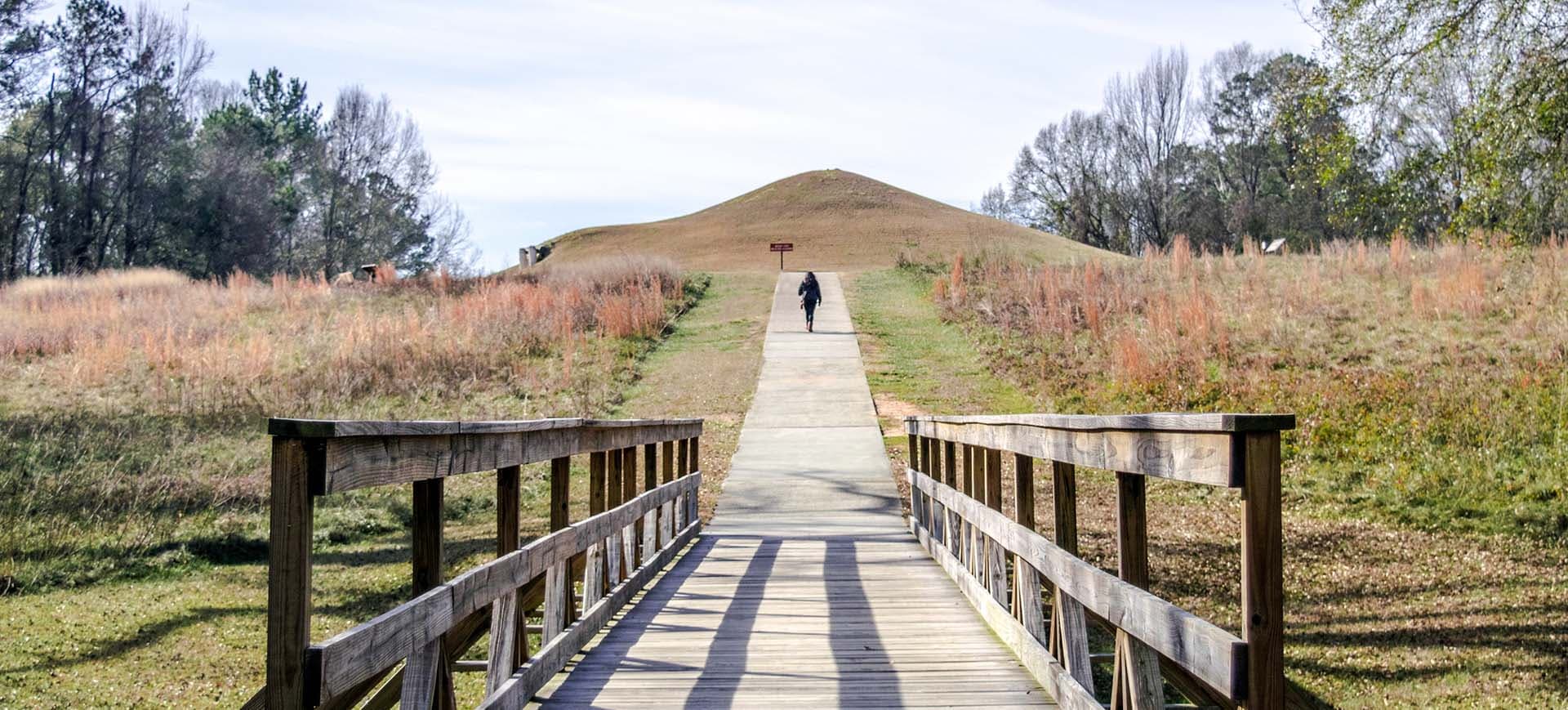 Walking path at the Ocmulgee Mounds National Historic Park with a wooden bridge and a green hill