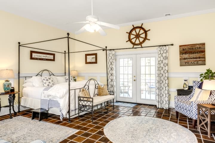 Spacious coastal-themed guest room featuring a four-poster bed, nautical decor, terracotta tile flooring, and French doors leading to a private outdoor area.