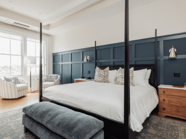 Elegant bedroom featuring a four-poster king bed, blue paneled accent wall, cozy seating, and ample natural light.